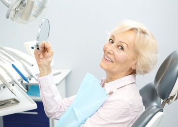 An older woman holding a handheld mirror and smiling after receiving her dentures