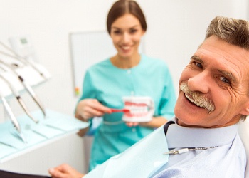 An older gentleman smiling in the dentist’s chair while a hygienist shows him how to properly care for his dentures in Crown Point