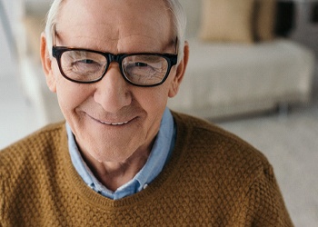 An older man wearing glasses and a sweater smiles after receiving his dental implant supported dentures in Crown Point