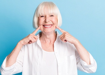 An older woman wearing white and pointing to her new smile after receiving dental implant supported dentures