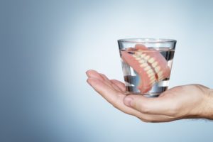 hand holding a glass of water with dentures soaking inside of it 