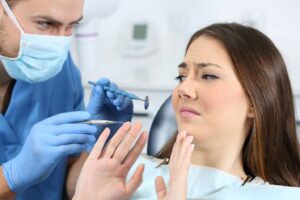 Worried dental patient holding up hands to approaching dentist with tools.